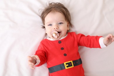 Cute baby wearing festive Christmas costume with pacifier on white bedsheet, top view