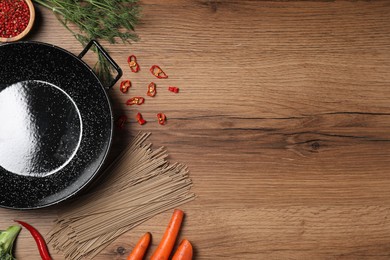 Photo of Empty iron wok surrounded by raw ingredients on wooden table, flat lay. Space for text