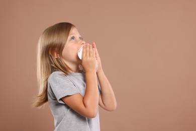 Photo of Suffering from allergy. Little girl with tissue sneezing on light brown background, space for text