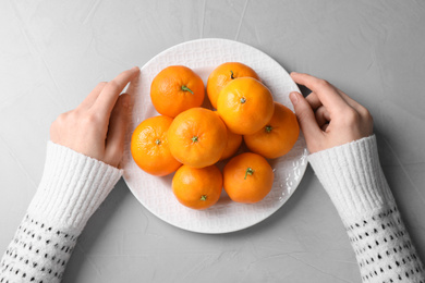 Photo of Woman with plate of fresh ripe tangerines at grey table, top view