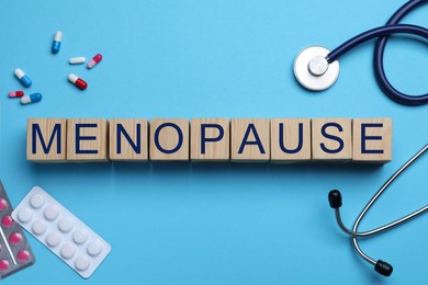 Wooden cubes with word Menopause, stethoscope and pills on light blue background, flat lay