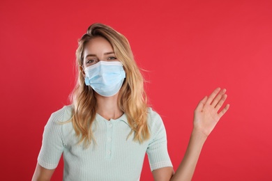 Photo of Woman in protective face mask showing hello gesture on red background. Keeping social distance during coronavirus pandemic