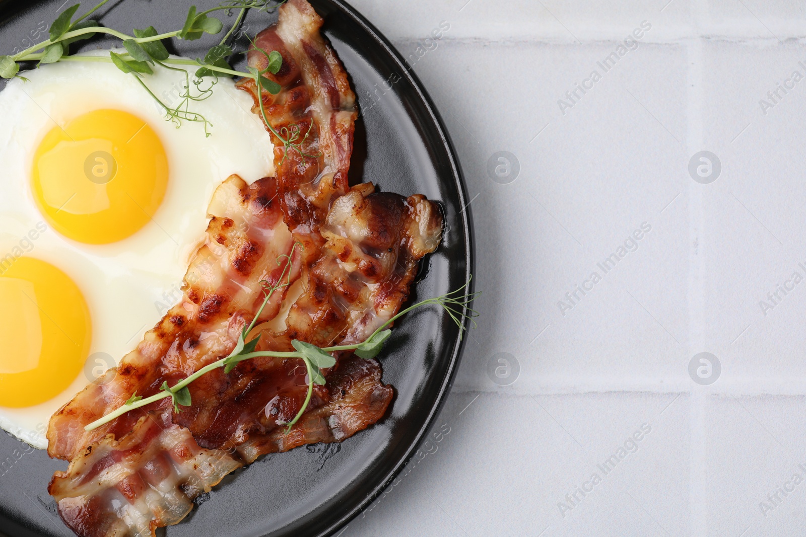 Photo of Fried eggs, bacon and microgreens on white tiled table, top view. Space for text