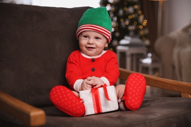 Photo of Cute little baby wearing Santa's elf clothes with Christmas gift in armchair at home