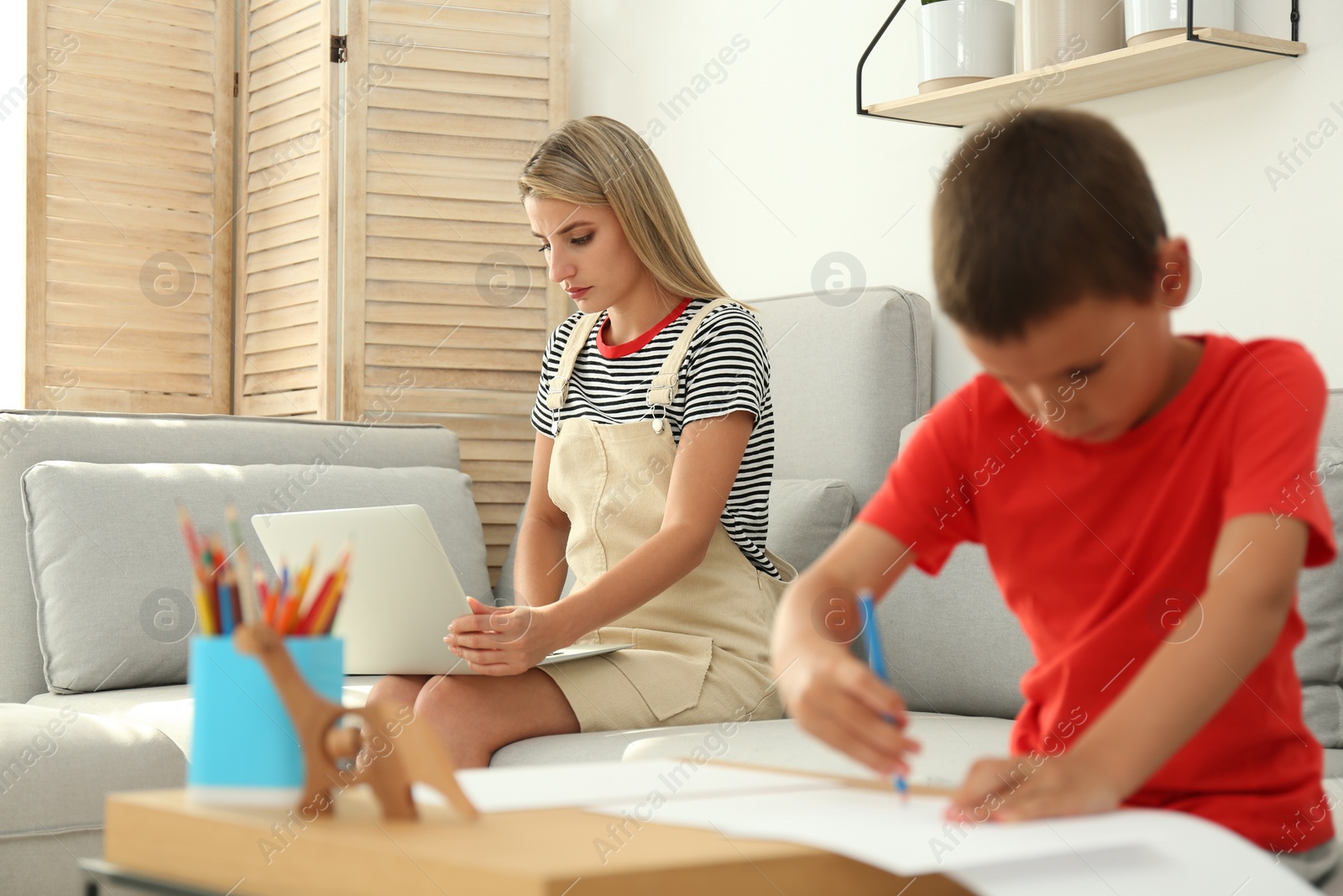 Photo of Young woman working on laptop while son drawing in living room. Home office concept