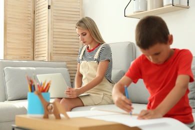 Photo of Young woman working on laptop while son drawing in living room. Home office concept