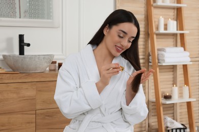 Photo of Happy young woman applying essential oil onto hair in bathroom