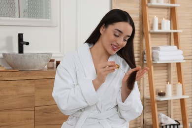 Happy young woman applying essential oil onto hair in bathroom