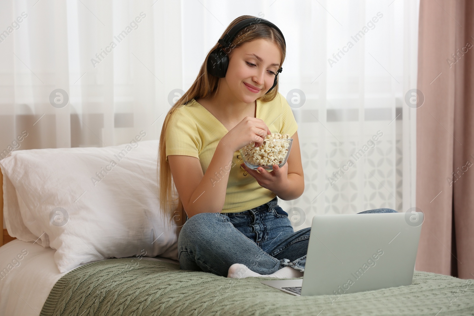 Photo of Teenage girl with headphones eating popcorn while using laptop on bed at home