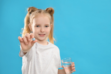 Photo of Little girl with vitamin pill and glass of water against blue background, focus on hand. Space for text