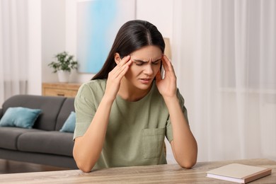 Photo of Sad woman suffering from headache at wooden table indoors