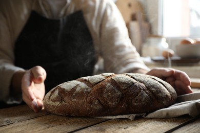 Photo of Man holding loaf of fresh bread at wooden table indoors, closeup