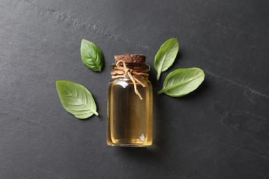 Photo of Bottle of essential basil oil and fresh leaves on dark grey table, flat lay