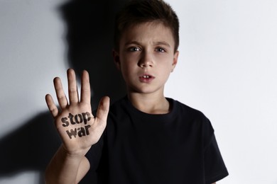 Image of Unhappy little boy showing palm with phrase Stop War near white wall