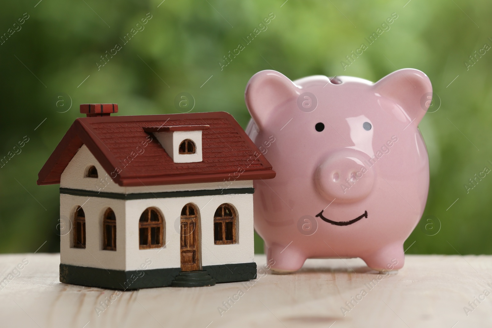 Photo of Piggy bank and little house toy on wooden table outdoors