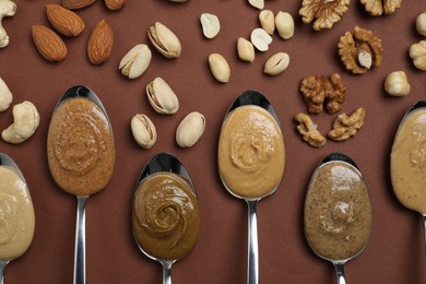 Tasty nut butters in spoons and raw nuts on brown table, flat lay