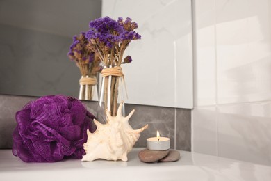 Photo of Purple shower puff, shell, burning candle and vase with flowers on sink in bathroom. Space for text