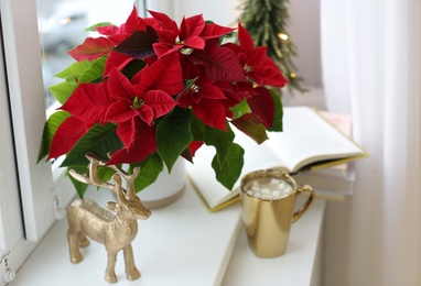 Photo of Beautiful poinsettia, cup of hot cocoa and decorative deer on window sill. Traditional Christmas flower