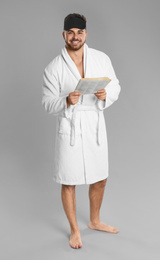 Young man in bathrobe reading newspaper on grey background