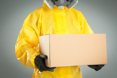 Photo of Man wearing chemical protective suit with cardboard box on light grey background, closeup. Prevention of virus spread