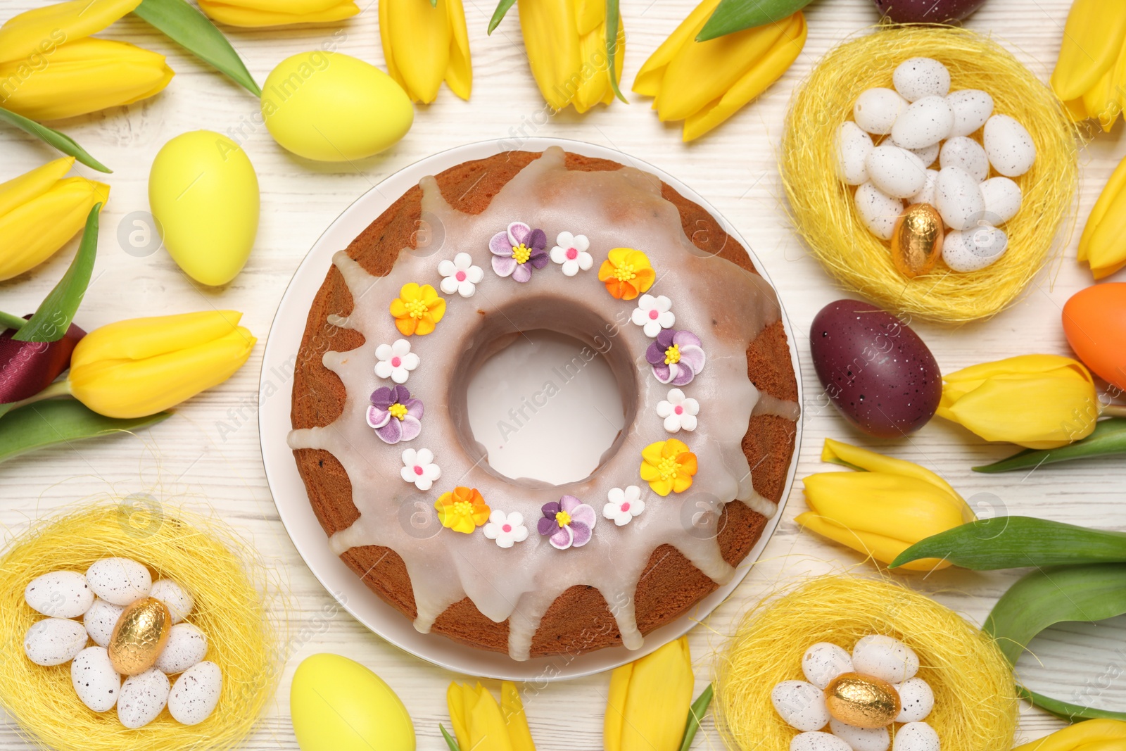 Photo of Flat lay composition of delicious Easter cake decorated with sprinkles, eggs and beautiful tulips on white wooden table