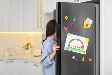 Photo of Woman opening refrigerator door with child's drawing, notes and magnets in kitchen