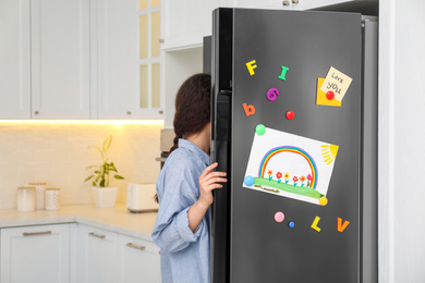 Woman opening refrigerator door with child's drawing, notes and magnets in kitchen