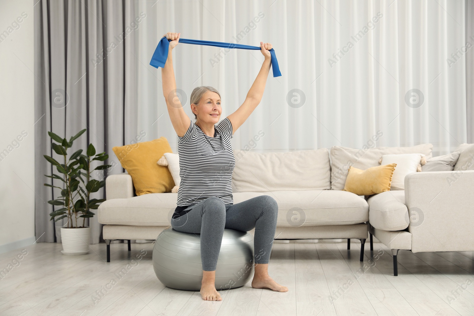 Photo of Senior woman doing exercise with elastic resistance band on fitness ball at home