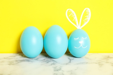 Image of One egg with drawn face and ears as Easter bunny among others on white marble table against yellow background