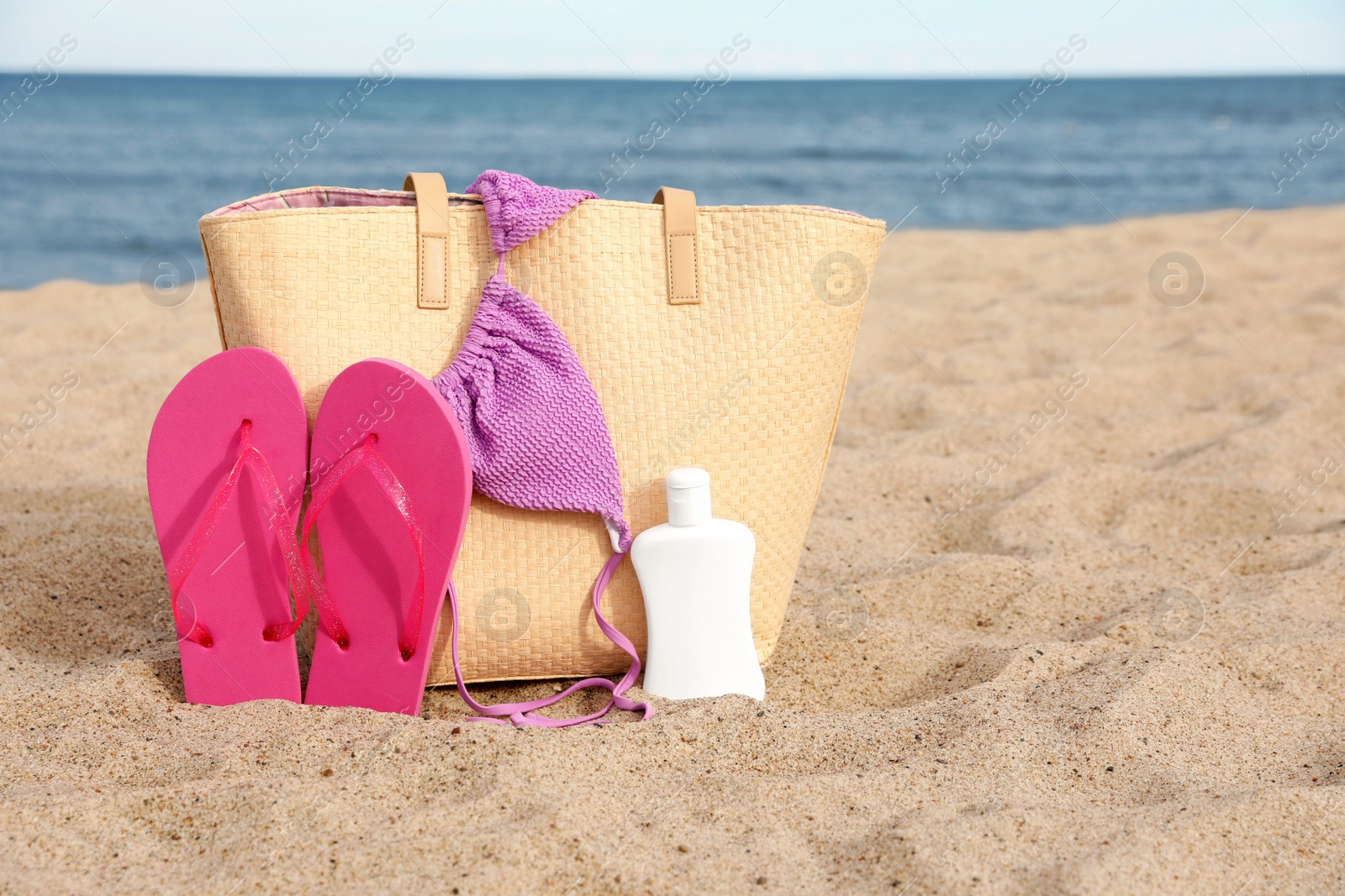 Photo of Summer bag with slippers, sunscreen and bikini top on sand near sea, space for text