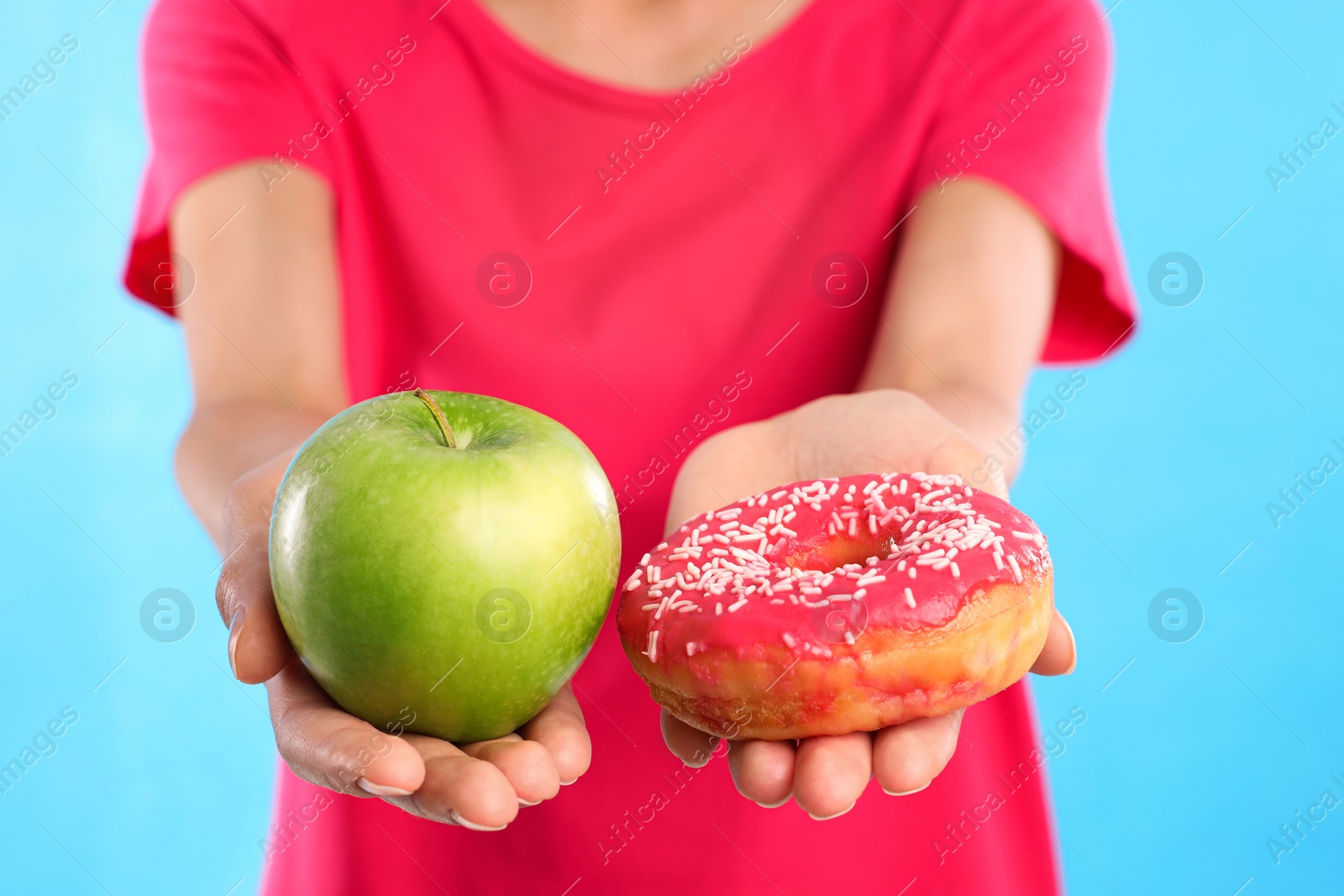 Photo of Woman choosing between doughnut and fresh apple on light blue background, closeup