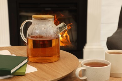 Photo of Teapot with cups of hot drink and books on wooden tables near decorative fireplace in room. Interior design