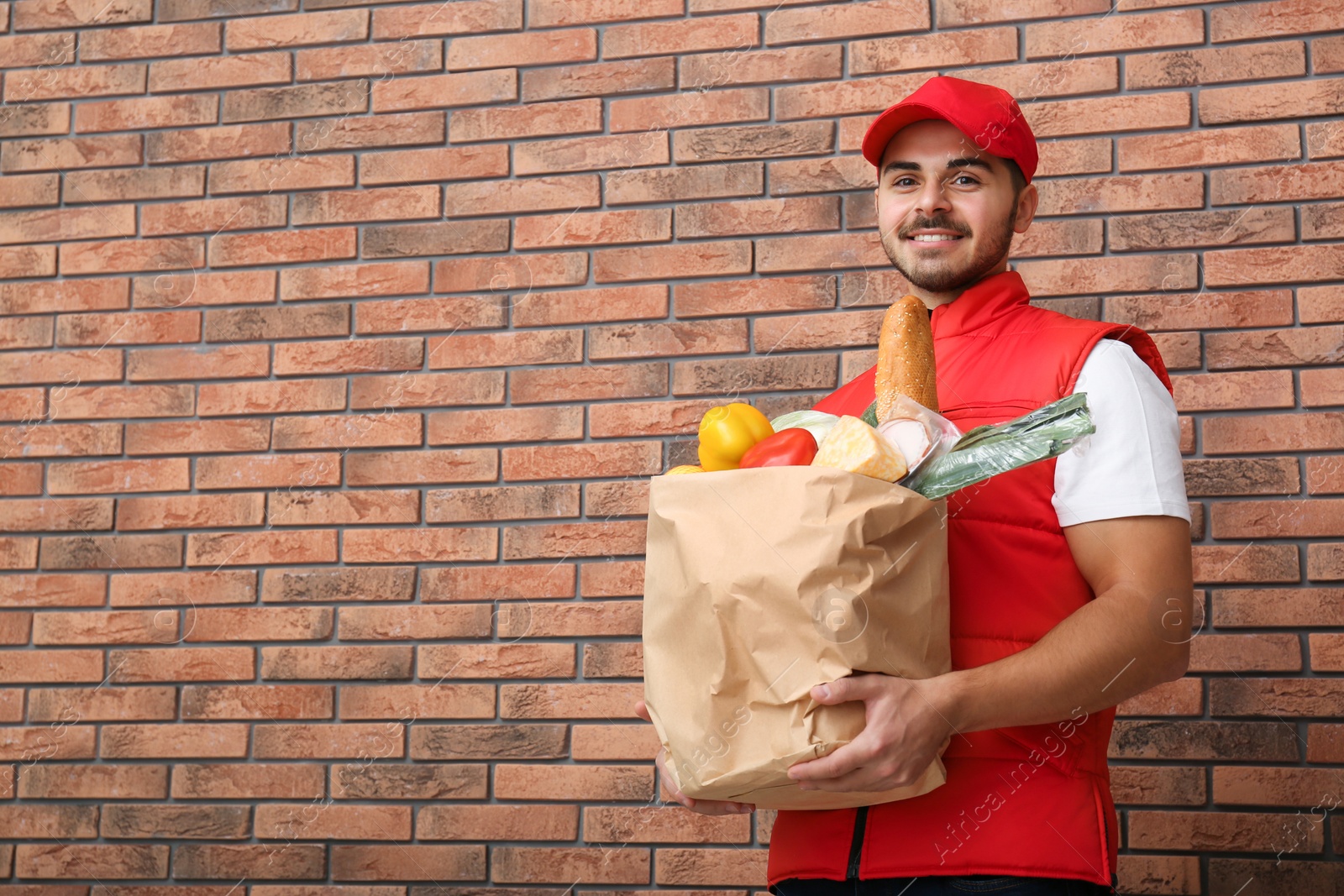 Photo of Food delivery courier holding paper bag with products near brick wall. Space for text