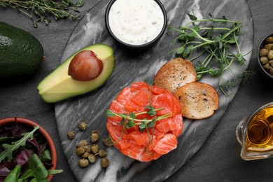 Photo of Delicious salmon tartare with avocado, sauce and croutons on dark table, flat lay