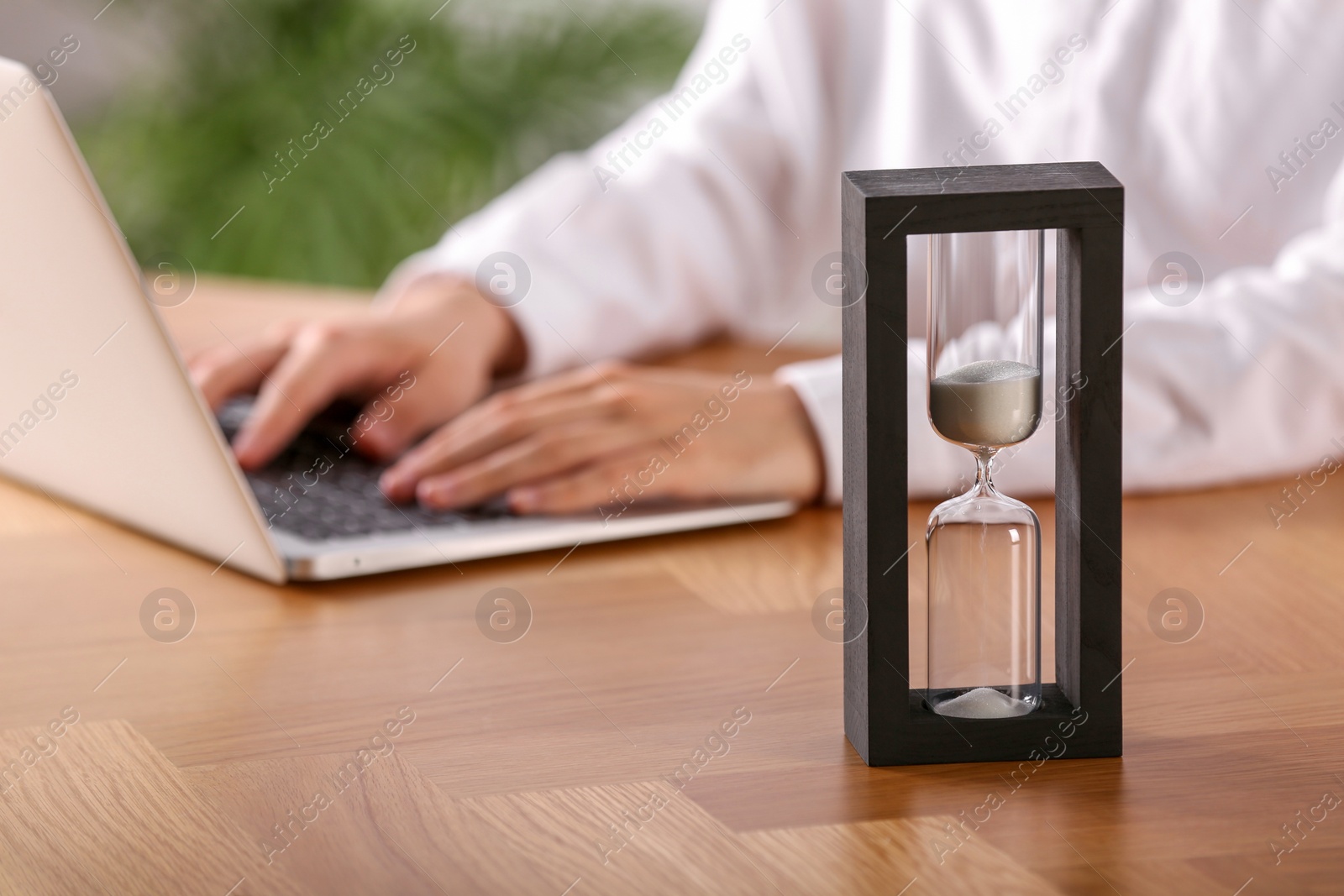 Photo of Hourglass with flowing sand on wooden table, selective focus. Man using laptop indoors