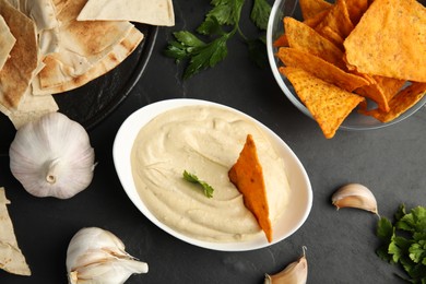 Photo of Delicious hummus with nachos and parsley on black table, flat lay