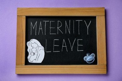 Chalkboard with phrase Maternity Leave, baby pacifier, paper cutout of mother and child on violet background, top view