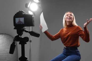 Photo of Casting call. Emotional woman with script sitting on chair and performing in front of camera against grey background in studio, selective focus