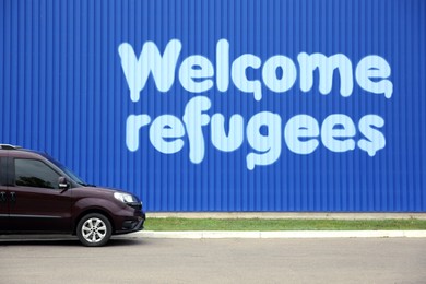 Car parked near blue wall with phrase WELCOME REFUGEES outdoors
