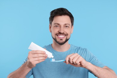 Photo of Happy man squeezing toothpaste from tube onto plastic toothbrush on light blue background