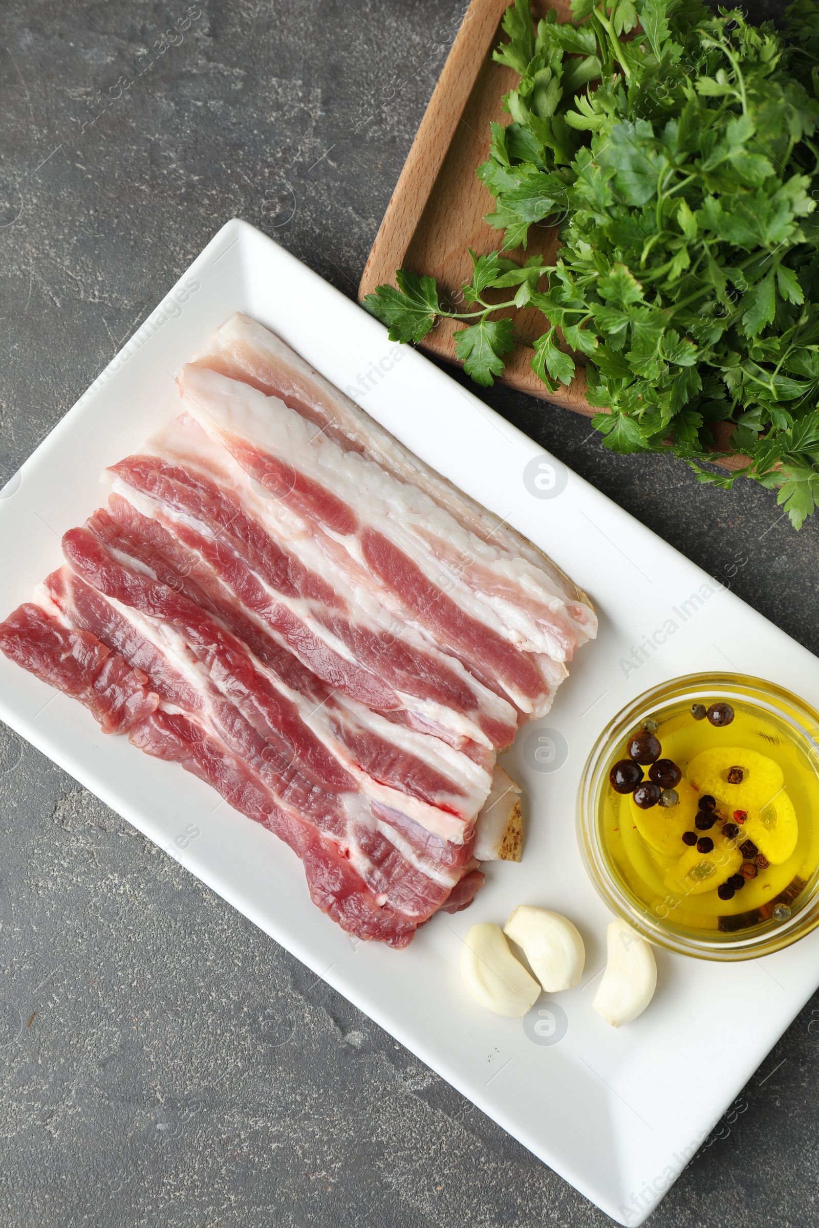 Photo of Pieces of raw pork belly, oil, parsley and garlic on grey textured table, top view