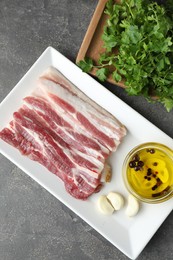 Pieces of raw pork belly, oil, parsley and garlic on grey textured table, top view