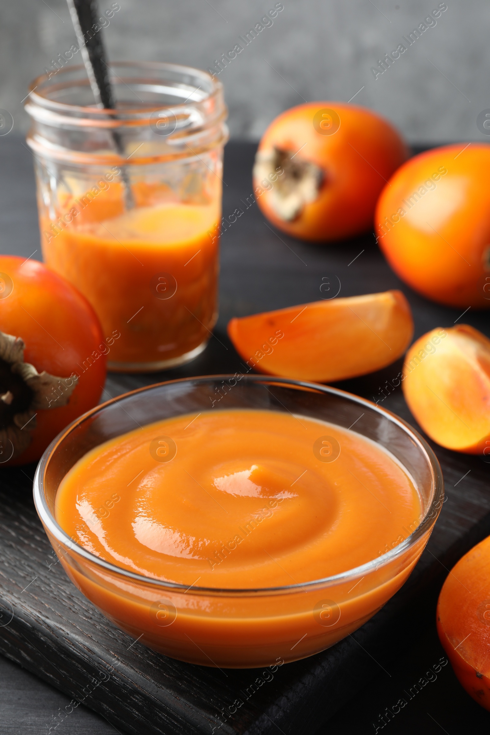 Photo of Delicious persimmon jam in glass dishware and fresh fruits on dark gray wooden table, closeup