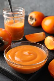 Delicious persimmon jam in glass dishware and fresh fruits on dark gray wooden table, closeup