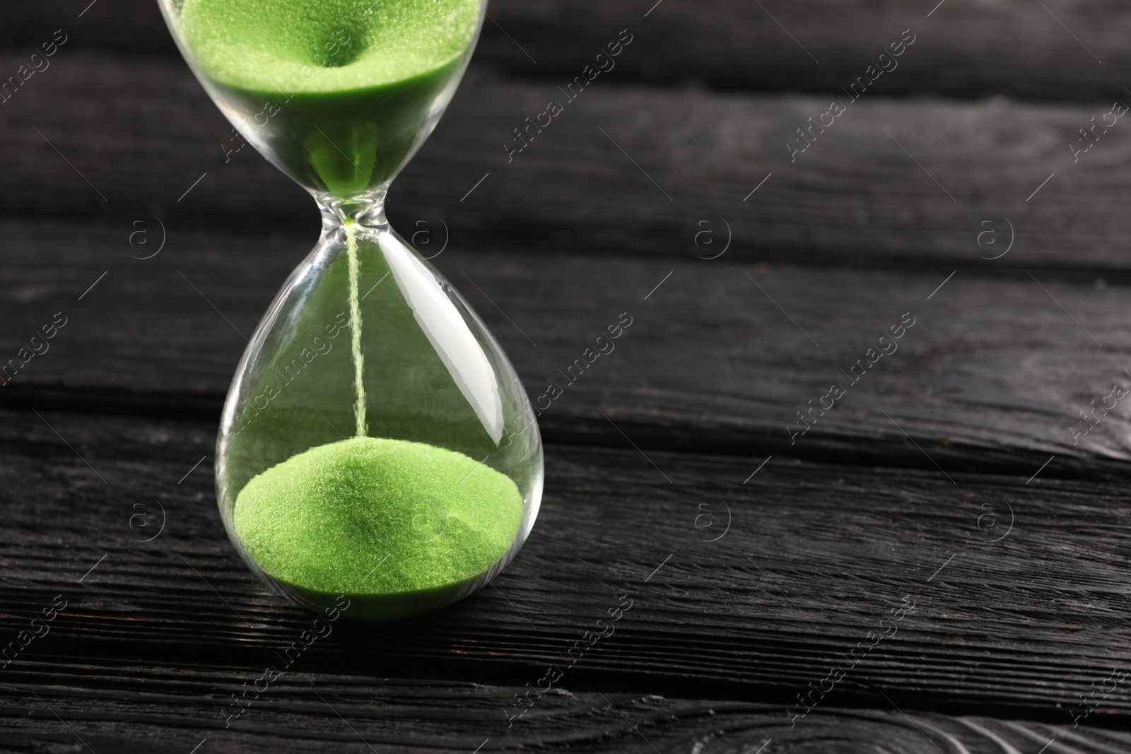 Photo of Hourglass with light green flowing sand on black wooden table, closeup. Space for text