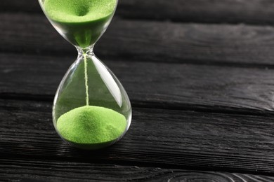 Hourglass with light green flowing sand on black wooden table, closeup. Space for text