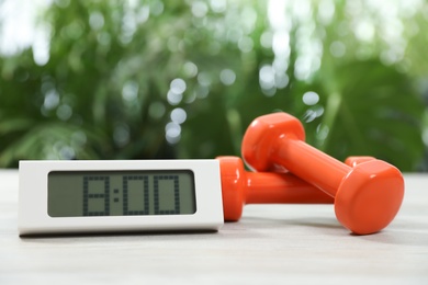 Photo of Digital alarm clock and dumbbells on white wooden table outdoors. Morning exercise