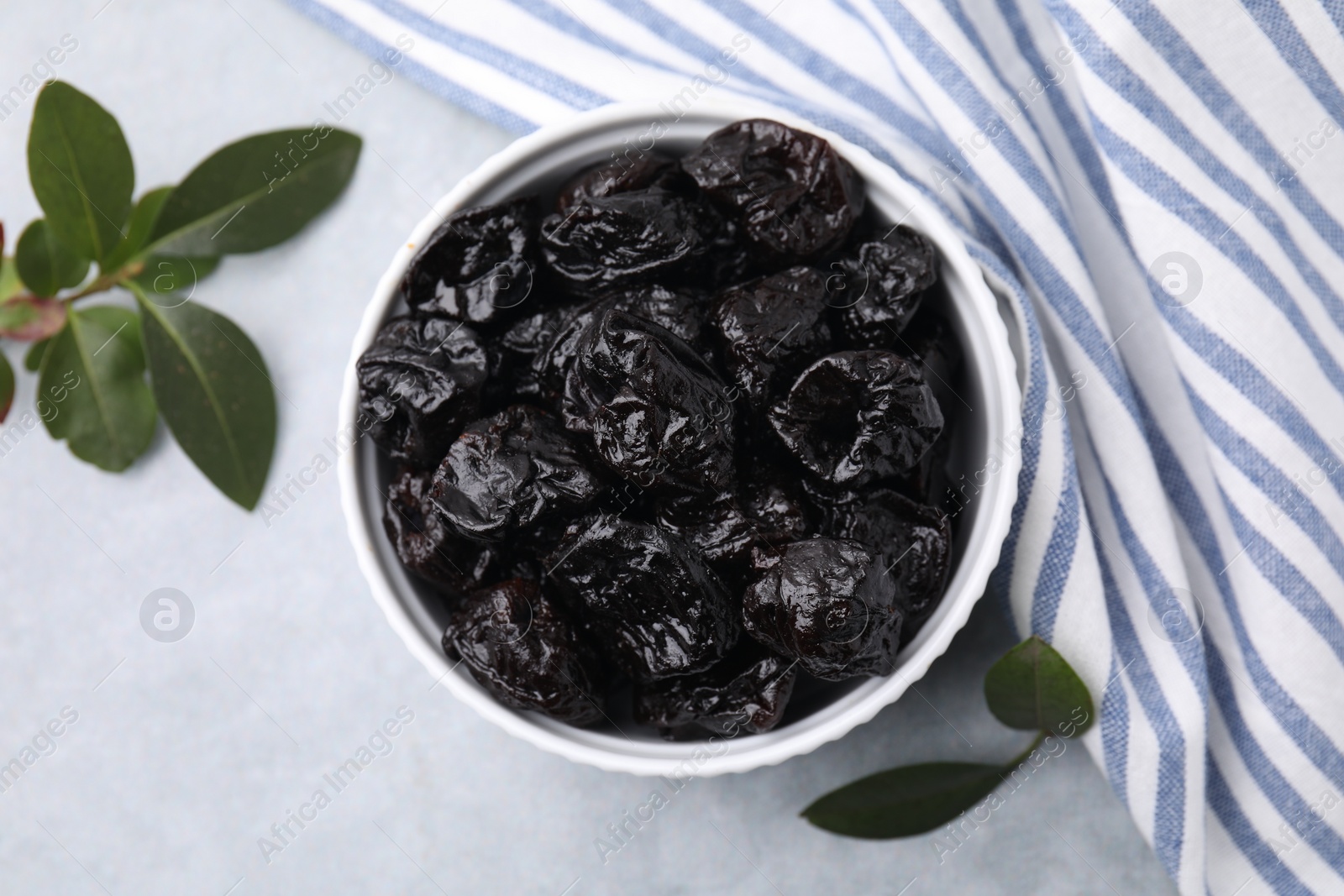 Photo of Sweet dried prunes in bowl and green leaves on light grey table, top view