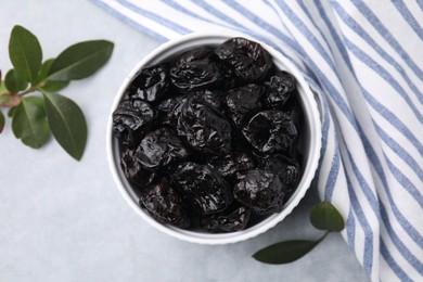 Sweet dried prunes in bowl and green leaves on light grey table, top view