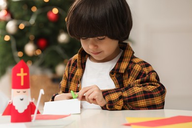 Cute little boy cutting paper at table with Saint Nicholas toy indoors